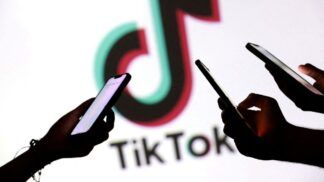 Imagem mostra as mãos de três pessoas mexendo no celular, em primeiro plano. No segundo plano, há o logo da empresa TikTok.