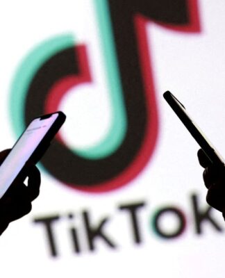 Imagem mostra as mãos de três pessoas mexendo no celular, em primeiro plano. No segundo plano, há o logo da empresa TikTok.