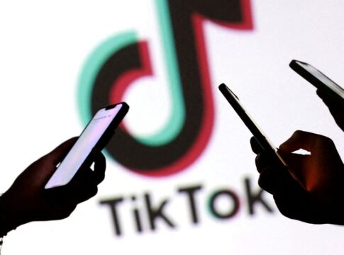 Imagem mostra as mãos de três pessoas mexendo no celular, em primeiro plano. No segundo plano, há o logo da empresa TikTok.