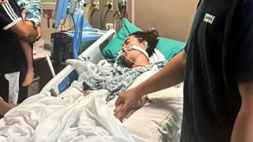 A imagem retrata uma mulher em estado grave na cama de um hospital, com o suporte de equipamentos médicos, enquanto familiares ou a acompanham, segurando na mão dela. A cama é branca com lençóis brancos, e a paciente está coberta por um cobertor estampado em tons de cinza. Vários fios, tubos e monitores estão visíveis ao redor da paciente e da cama, característicos de uma unidade de terapia intensiva ou cuidados críticos.