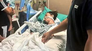 A imagem retrata uma mulher em estado grave na cama de um hospital, com o suporte de equipamentos médicos, enquanto familiares ou a acompanham, segurando na mão dela. A cama é branca com lençóis brancos, e a paciente está coberta por um cobertor estampado em tons de cinza. Vários fios, tubos e monitores estão visíveis ao redor da paciente e da cama, característicos de uma unidade de terapia intensiva ou cuidados críticos.