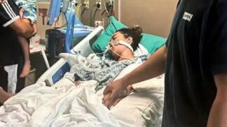 A imagem retrata uma mulher em estado grave na cama de um hospital, com o suporte de equipamentos médicos, enquanto familiares ou a acompanham, segurando na mão dela. A cama é branca com lençóis brancos, e a paciente está coberta por um cobertor estampado em tons de cinza. Vários fios, tubos e monitores estão visíveis ao redor da paciente e da cama, característicos de uma unidade de terapia intensiva ou cuidados críticos.