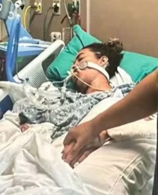 A imagem retrata uma mulher em estado grave na cama de um hospital, com o suporte de equipamentos médicos, enquanto familiares ou a acompanham, segurando na mão dela. A cama é branca com lençóis brancos, e a paciente está coberta por um cobertor estampado em tons de cinza. Vários fios, tubos e monitores estão visíveis ao redor da paciente e da cama, característicos de uma unidade de terapia intensiva ou cuidados críticos.