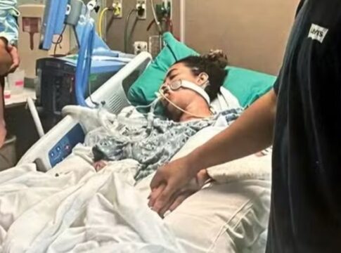 A imagem retrata uma mulher em estado grave na cama de um hospital, com o suporte de equipamentos médicos, enquanto familiares ou a acompanham, segurando na mão dela. A cama é branca com lençóis brancos, e a paciente está coberta por um cobertor estampado em tons de cinza. Vários fios, tubos e monitores estão visíveis ao redor da paciente e da cama, característicos de uma unidade de terapia intensiva ou cuidados críticos.