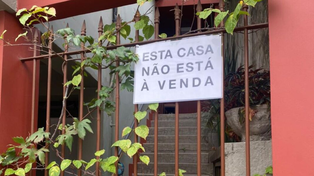 Uma foto de um portão de ferro, coberto por algumas trepadeiras de folhas verdes, que revela a entrada de uma casa. O portão está pintado de marrom, e a parede, ao seu lado direito, é de um tom de vermelho terracota. Pendurado no portão, no centro da imagem, há um cartaz retangular branco com letras pretas em caixa alta, que diz: 