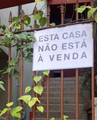 Uma foto de um portão de ferro, coberto por algumas trepadeiras de folhas verdes, que revela a entrada de uma casa. O portão está pintado de marrom, e a parede, ao seu lado direito, é de um tom de vermelho terracota. Pendurado no portão, no centro da imagem, há um cartaz retangular branco com letras pretas em caixa alta, que diz: 