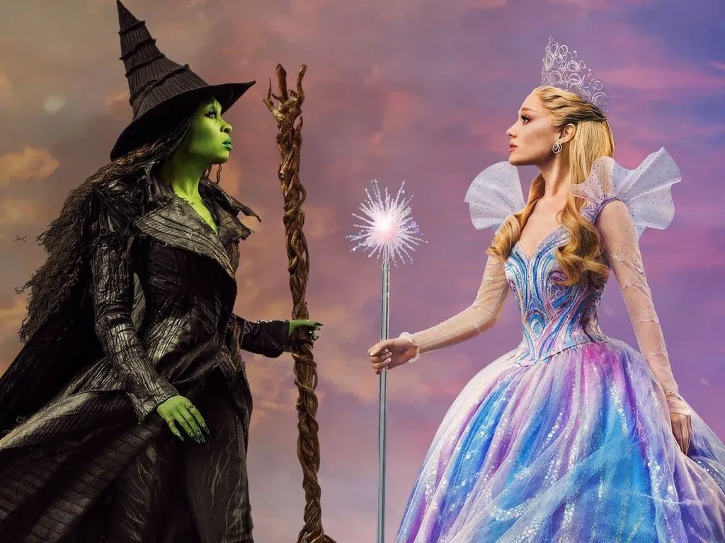 Duas mulheres se encaram em um fundo de céu roxo. Uma mulher segura uma vassoura e é verde. A outra é branca e segura uma varinha.