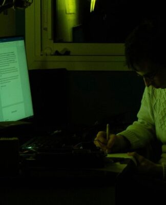 Homem no escuro, com a tela do computador ligada, escrevendo em um caderno à mão