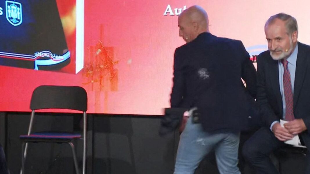 Luis Rubiales esquivando de uma ovada; a parede ao fundo já mostra pelo menos dois ovos quebrados