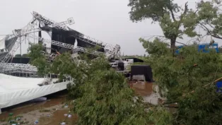 Estrutura metálica de palco desabada após tempestade em área aberta, com árvores caídas, lonas danificadas e o chão coberto de lama e destroços.