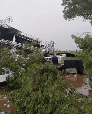 Estrutura metálica de palco desabada após tempestade em área aberta, com árvores caídas, lonas danificadas e o chão coberto de lama e destroços.
