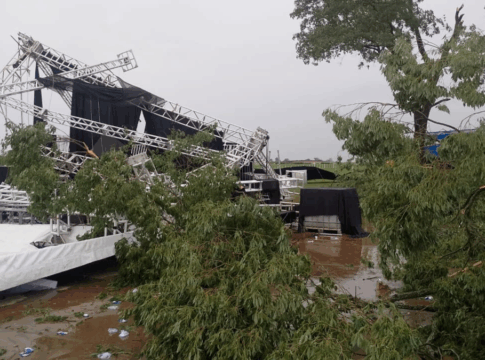 Estrutura metálica de palco desabada após tempestade em área aberta, com árvores caídas, lonas danificadas e o chão coberto de lama e destroços.