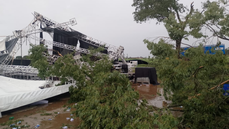 Estrutura metálica de palco desabada após tempestade em área aberta, com árvores caídas, lonas danificadas e o chão coberto de lama e destroços.