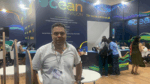 Homem usando camisa branca e crachá posa em pé diante do estande “Ocean Pavilion”, onde várias pessoas trabalham e conversam ao fundo; o espaço tem paredes decoradas com tons de azul e verde e a frase “The ocean unites us all.”