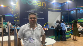 Homem usando camisa branca e crachá posa em pé diante do estande “Ocean Pavilion”, onde várias pessoas trabalham e conversam ao fundo; o espaço tem paredes decoradas com tons de azul e verde e a frase “The ocean unites us all.”