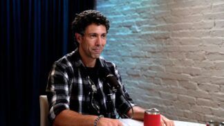 Rodrigo Oliveira sentado em uma mesa, ao lado de uma parede de tijolos brancos