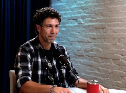 Rodrigo Oliveira sentado em uma mesa, ao lado de uma parede de tijolos brancos