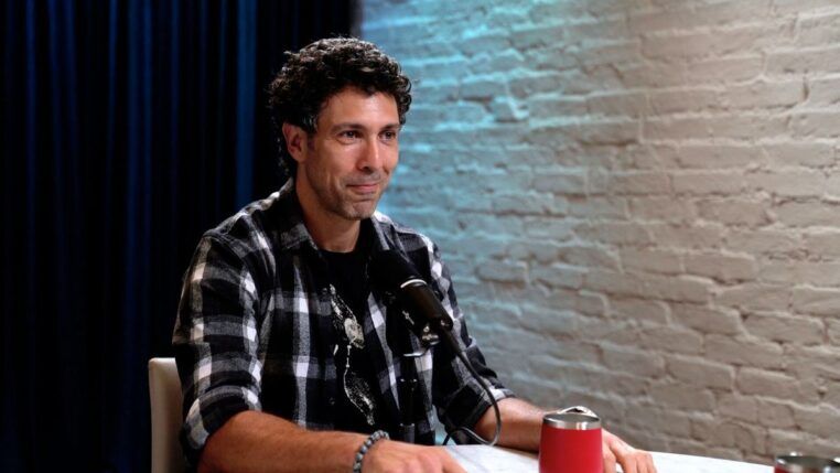 Rodrigo Oliveira sentado em uma mesa, ao lado de uma parede de tijolos brancos