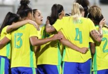 Jogadoras da seleção feminina de futebol do Brasil, vestindo uniformes amarelos e azuis, se abraçam em roda durante um momento de união antes da partida.