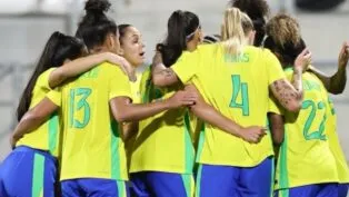 Jogadoras da seleção feminina de futebol do Brasil, vestindo uniformes amarelos e azuis, se abraçam em roda durante um momento de união antes da partida.