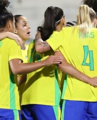 Jogadoras da seleção feminina de futebol do Brasil, vestindo uniformes amarelos e azuis, se abraçam em roda durante um momento de união antes da partida.