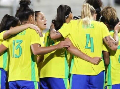 Jogadoras da seleção feminina de futebol do Brasil, vestindo uniformes amarelos e azuis, se abraçam em roda durante um momento de união antes da partida.