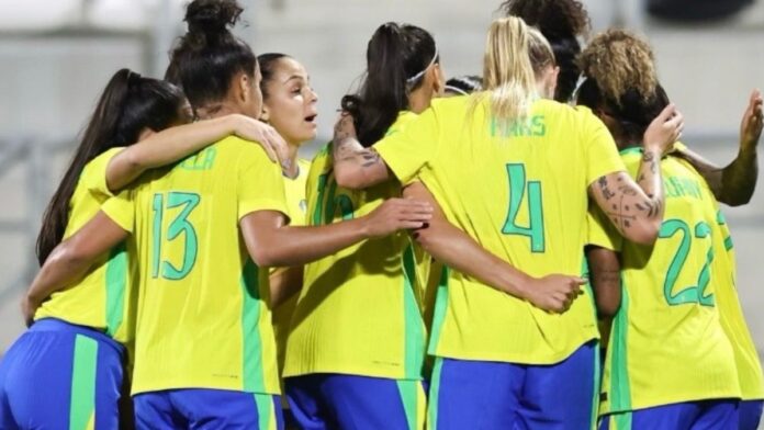 Jogadoras da seleção feminina de futebol do Brasil, vestindo uniformes amarelos e azuis, se abraçam em roda durante um momento de união antes da partida.