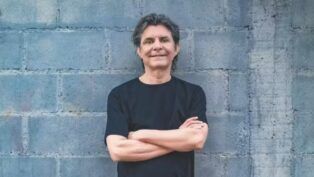 Lô Borges encostado em uma parede de tijolos cinza