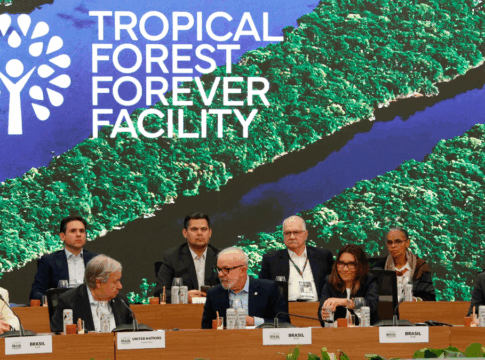 Autoridades e representantes de diferentes países e organizações sentados à mesa durante evento do “Tropical Forest Forever Facility”, com imagem aérea de floresta tropical e rios projetada ao fundo.