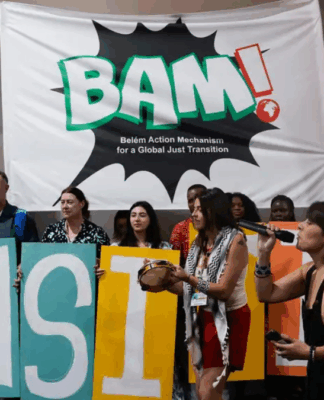 Grupo de ativistas segura letras coloridas formando a palavra “TRANSITION” em frente a um grande banner com a inscrição “BAM! Belém Action Mechanism for a Global Just Transition”, enquanto faixas verticais trazem palavras como “Care”, “Wisdom”, “Inclusion” e “Labour Rights” em um ato dentro de espaço fechado.