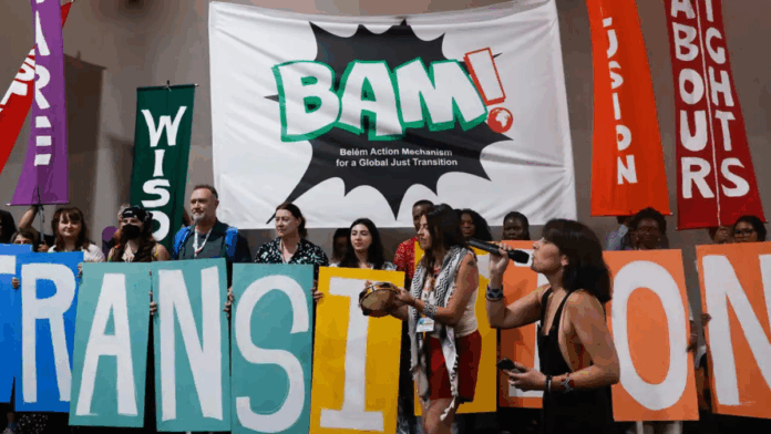 Grupo de ativistas segura letras coloridas formando a palavra “TRANSITION” em frente a um grande banner com a inscrição “BAM! Belém Action Mechanism for a Global Just Transition”, enquanto faixas verticais trazem palavras como “Care”, “Wisdom”, “Inclusion” e “Labour Rights” em um ato dentro de espaço fechado.