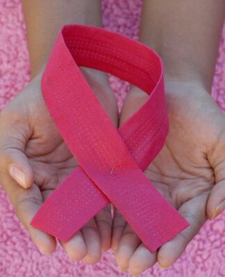 Mãos em concha seguram um laço rosa, símbolo de conscientização sobre o câncer de mama, sobre um fundo felpudo também rosa.