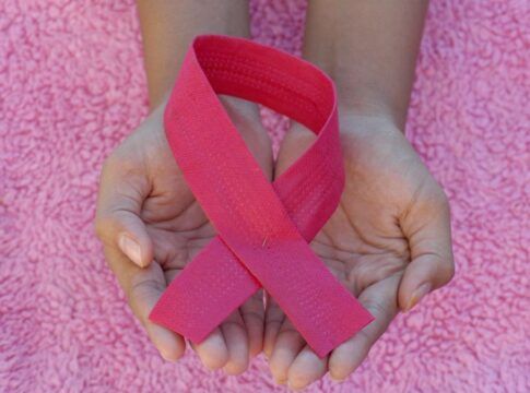 Mãos em concha seguram um laço rosa, símbolo de conscientização sobre o câncer de mama, sobre um fundo felpudo também rosa.