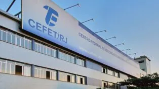 Fachada do prédio do CEFET/RJ, com placas institucionais visíveis, janelas amplas e bandeiras hasteadas, cercado por palmeiras e carros estacionados em frente.