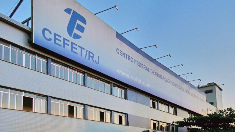 Fachada do prédio do CEFET/RJ, com placas institucionais visíveis, janelas amplas e bandeiras hasteadas, cercado por palmeiras e carros estacionados em frente.