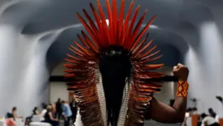 Pessoa indígena vista de costas, usando grande cocar de penas vermelhas e pulseira colorida, ergue o punho direito em gesto de resistência dentro de um salão moderno onde outras pessoas conversam ao fundo.