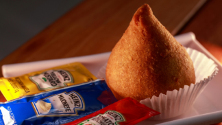 Coxinha dourada colocada em forminha de papel branco sobre bandeja, acompanhada de sachês de maionese, mostarda e ketchup da marca Heinz.