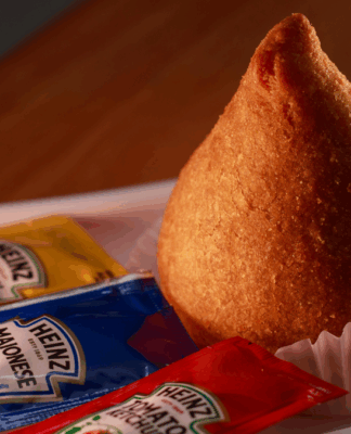 Coxinha dourada colocada em forminha de papel branco sobre bandeja, acompanhada de sachês de maionese, mostarda e ketchup da marca Heinz.