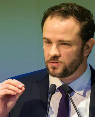 Homem de terno escuro e gravata roxa fala ao microfone durante um discurso, gesticulando com a mão levantada diante de um fundo em tom esverdeado.