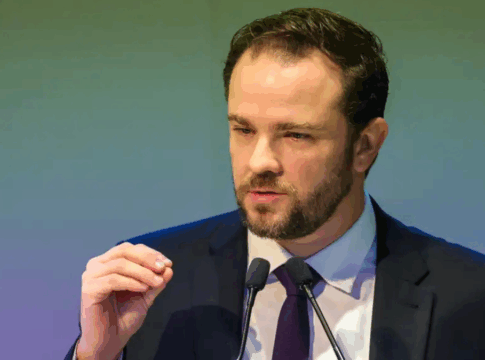 Homem de terno escuro e gravata roxa fala ao microfone durante um discurso, gesticulando com a mão levantada diante de um fundo em tom esverdeado.