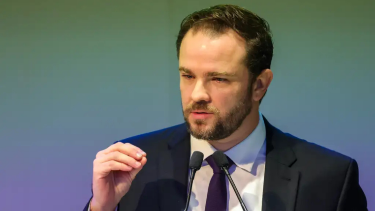 Homem de terno escuro e gravata roxa fala ao microfone durante um discurso, gesticulando com a mão levantada diante de um fundo em tom esverdeado.