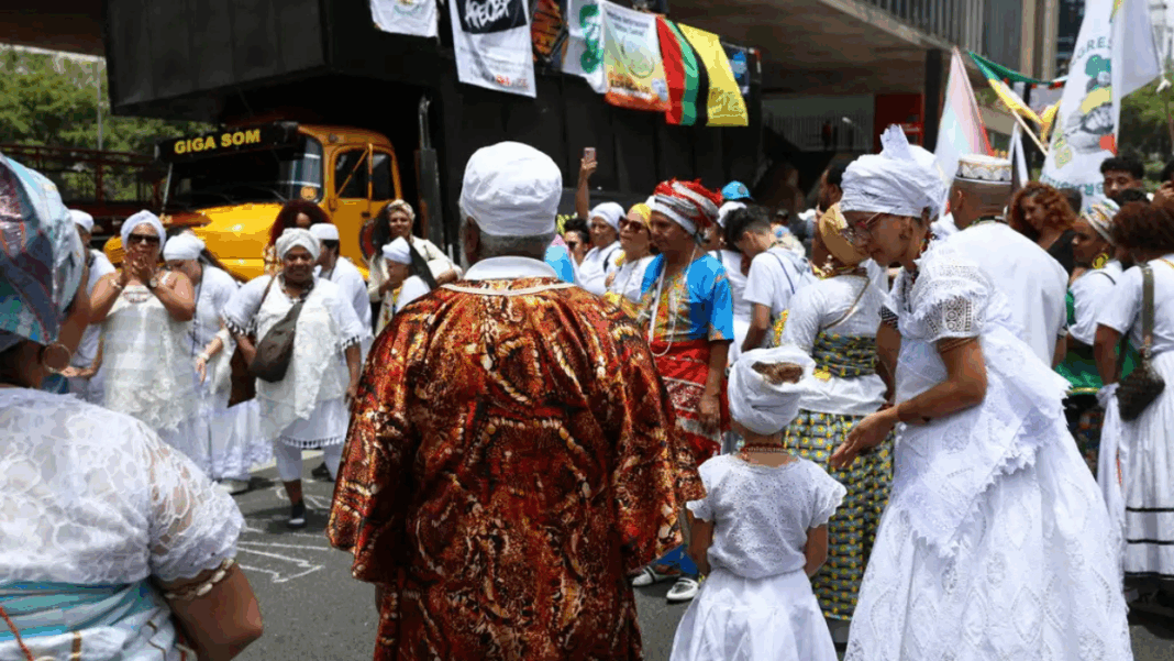 Grupo de pessoas vestidas com roupas tradicionais de matriz africana participa de celebração ao ar livre; muitas usam roupas brancas, turbantes e colares, enquanto outras vestem trajes coloridos. Banners e um caminhão de som aparecem ao fundo.