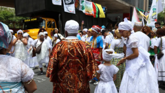 Grupo de pessoas vestidas com roupas tradicionais de matriz africana participa de celebração ao ar livre; muitas usam roupas brancas, turbantes e colares, enquanto outras vestem trajes coloridos. Banners e um caminhão de som aparecem ao fundo.