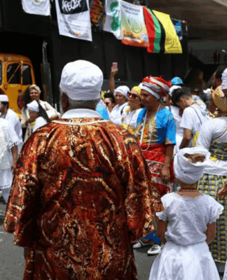 Grupo de pessoas vestidas com roupas tradicionais de matriz africana participa de celebração ao ar livre; muitas usam roupas brancas, turbantes e colares, enquanto outras vestem trajes coloridos. Banners e um caminhão de som aparecem ao fundo.