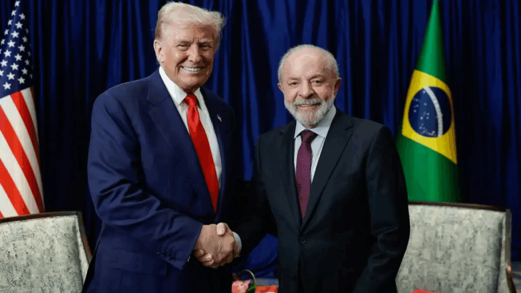 Dois homens de terno posam sorrindo enquanto se cumprimentam com um aperto de mão. Atrás deles aparecem as bandeiras dos Estados Unidos e do Brasil, com cortinas azuis ao fundo. À esquerda está o presidente americano, Donald Trump, e, à direita, o presidente brasileiro, Lula da Silva.
