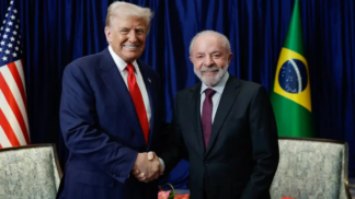 Dois homens de terno posam sorrindo enquanto se cumprimentam com um aperto de mão. Atrás deles aparecem as bandeiras dos Estados Unidos e do Brasil, com cortinas azuis ao fundo. À esquerda está o presidente americano, Donald Trump, e, à direita, o presidente brasileiro, Lula da Silva.