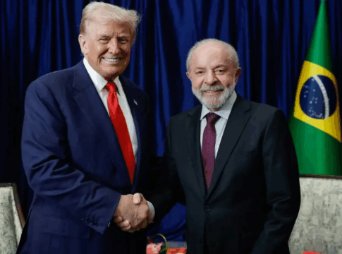 Dois homens de terno posam sorrindo enquanto se cumprimentam com um aperto de mão. Atrás deles aparecem as bandeiras dos Estados Unidos e do Brasil, com cortinas azuis ao fundo. À esquerda está o presidente americano, Donald Trump, e, à direita, o presidente brasileiro, Lula da Silva.