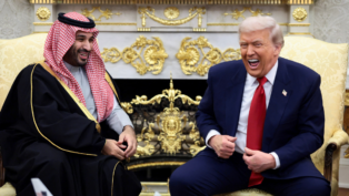 Dois homens sentados em poltronas claras conversam e riem em uma sala ornamentada com detalhes dourados; um deles veste terno azul com gravata vermelha, e o outro usa túnica tradicional preta com detalhes dourados e um lenço estampado vermelho e branco na cabeça.