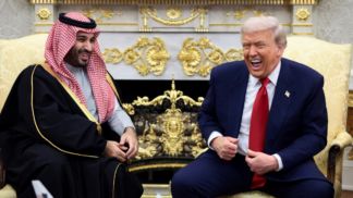 Dois homens sentados em poltronas claras conversam e riem em uma sala ornamentada com detalhes dourados; um deles veste terno azul com gravata vermelha, e o outro usa túnica tradicional preta com detalhes dourados e um lenço estampado vermelho e branco na cabeça.