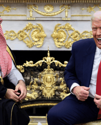 Dois homens sentados em poltronas claras conversam e riem em uma sala ornamentada com detalhes dourados; um deles veste terno azul com gravata vermelha, e o outro usa túnica tradicional preta com detalhes dourados e um lenço estampado vermelho e branco na cabeça.